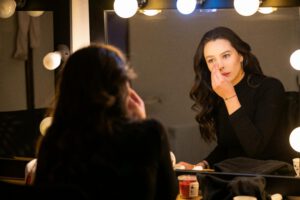 Young woman applies makeup in a brightly lit dressing room mirror, creating a focused and elegant atmosphere.
