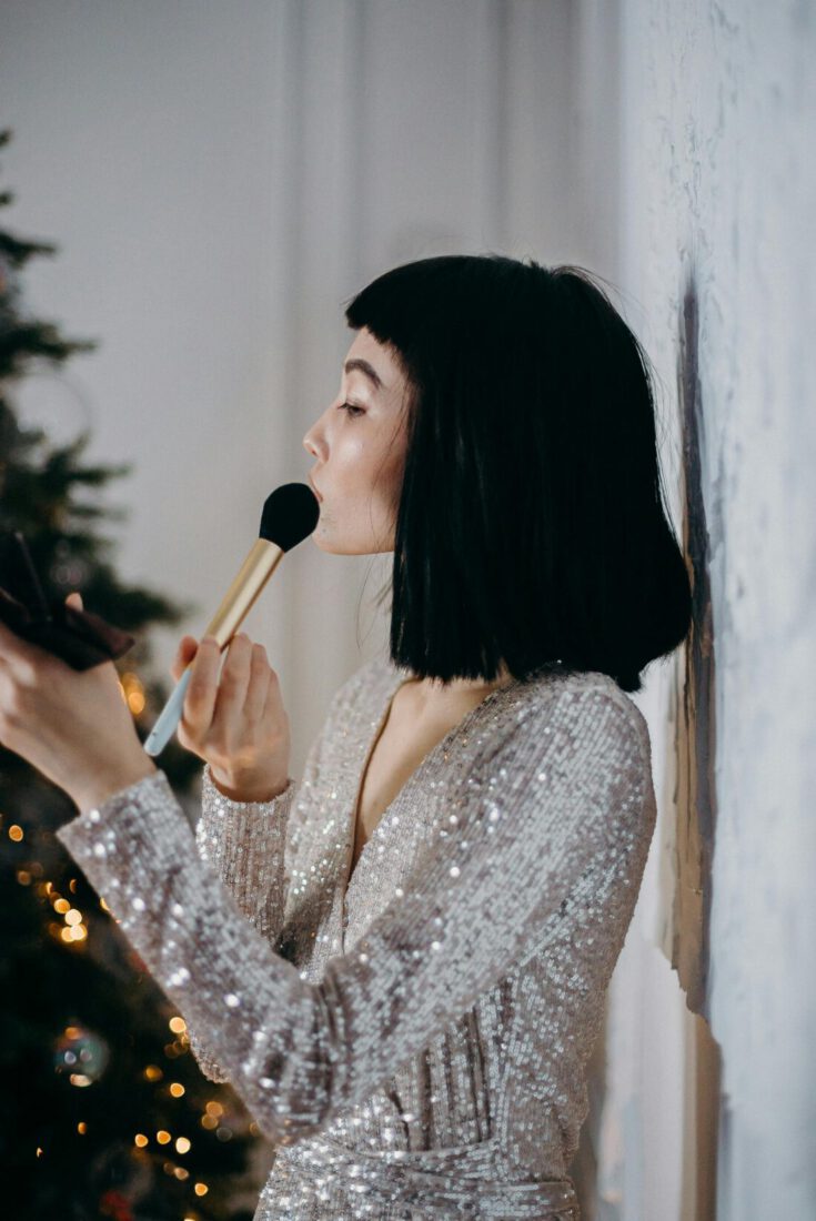 A woman in a silver dress applies makeup indoors beside a decorated Christmas tree.