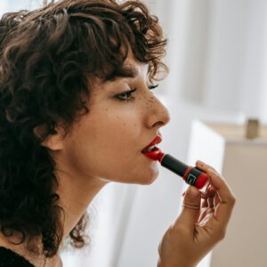 A young woman in profile view applies red lipstick indoors, embodying elegance and style.