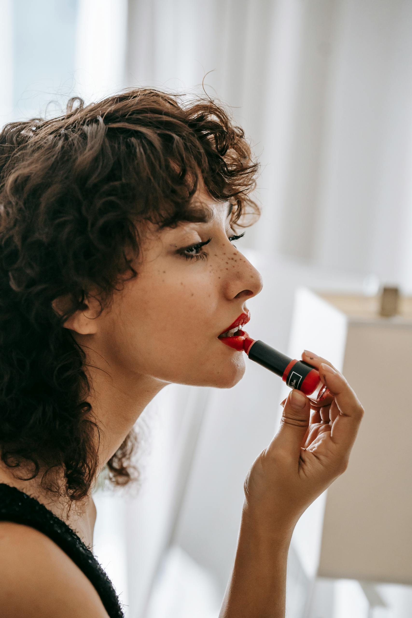 A young woman in profile view applies red lipstick indoors, embodying elegance and style.
