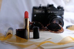 A close-up of a lipstick, vintage camera, and film roll against a soft backdrop.