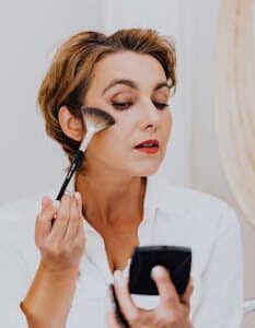 Woman using makeup brush in a well-lit indoor setting, enhancing her facial features.